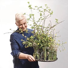 Foodbloggerin Heike Mueller traegt kurze Haare und haelt ein Tablett mit vollen Blumenvasen in den Haenden