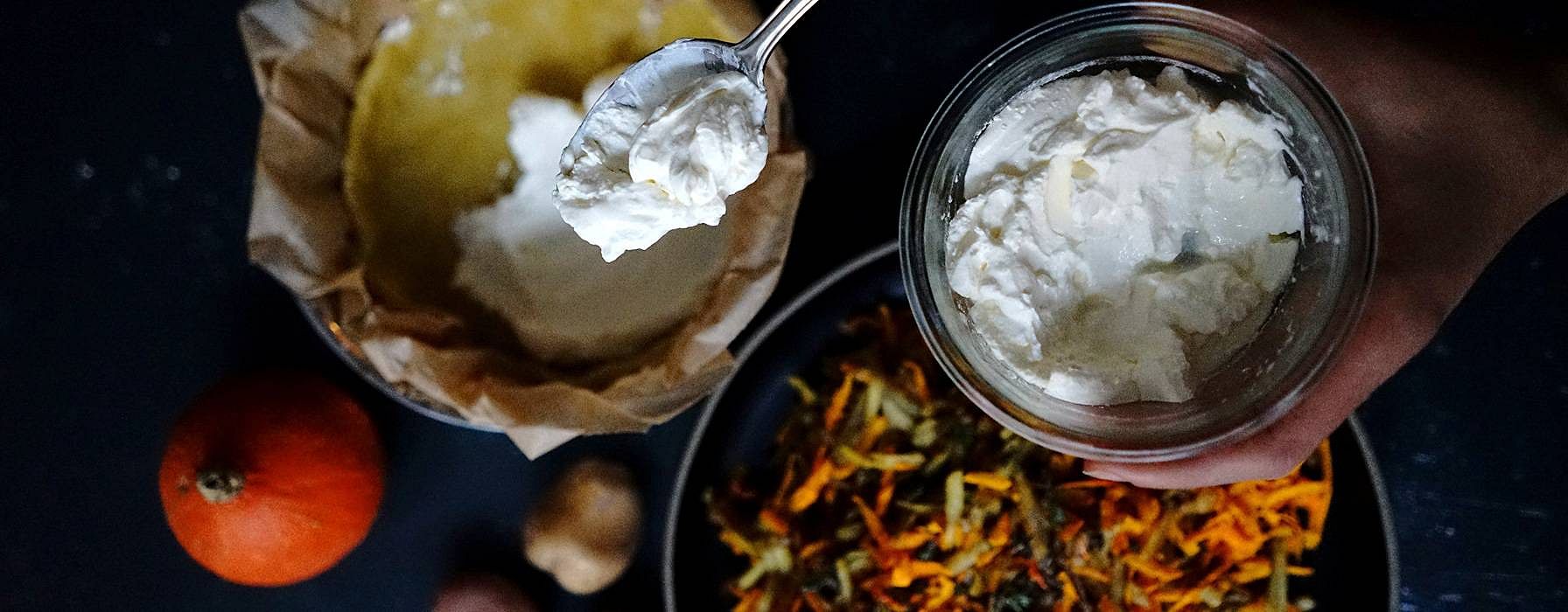 Hand haelt eine Glasschale mit Sauerrahm und einen Loeffel, darunter vorbereitete Kuchenform