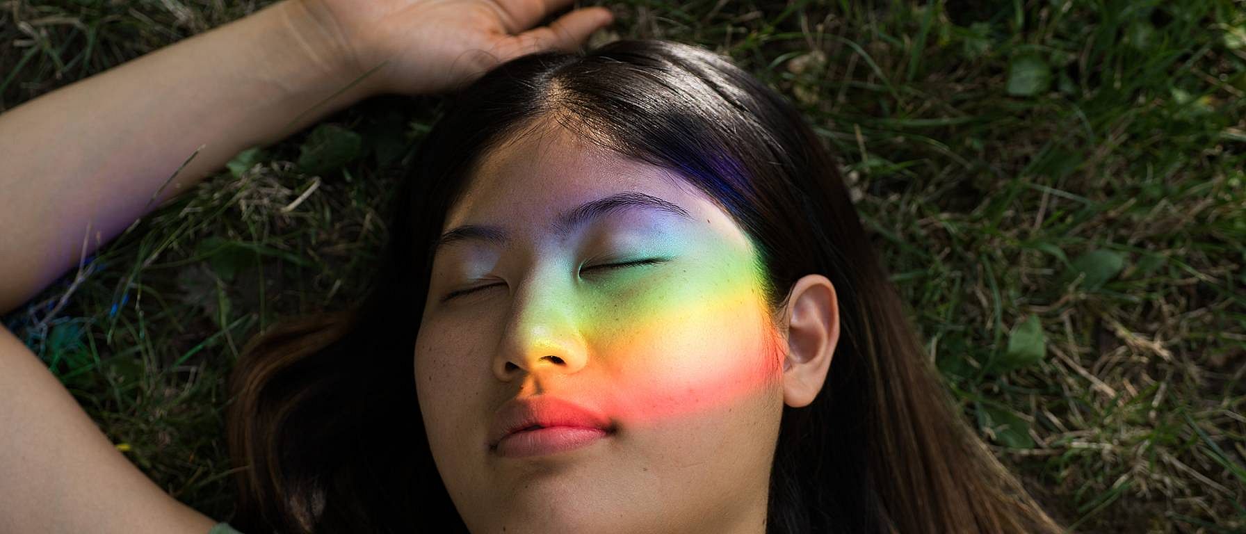 Frau mit geschlossenen Augen, im Gesicht die Spiegelung eines Regenbogenprismas