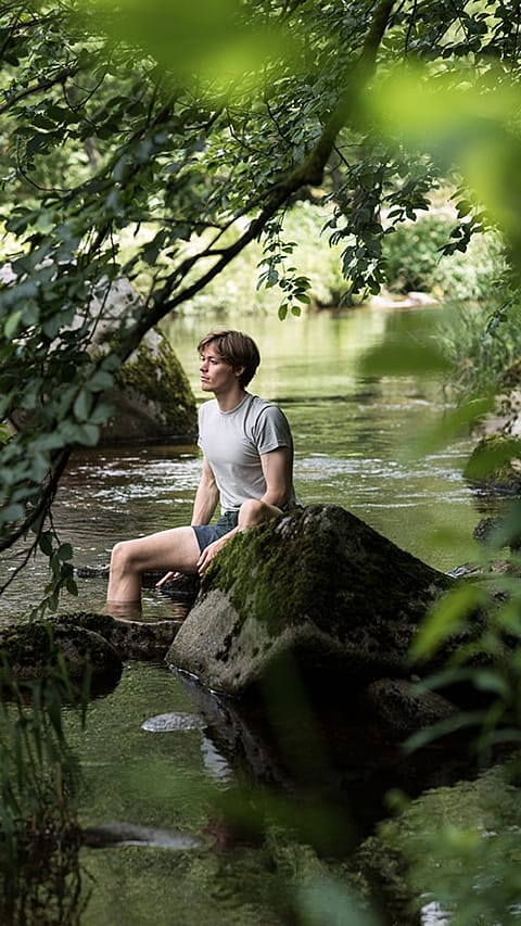 junger Mann haengt die Fuesse in einen Bach