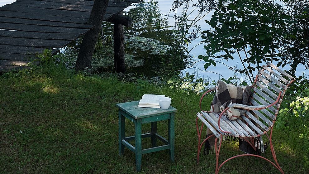 Stuhl mit Decke in der Natur an einem See, auf einem Tischchen stehen ein Buch und eine Tasse